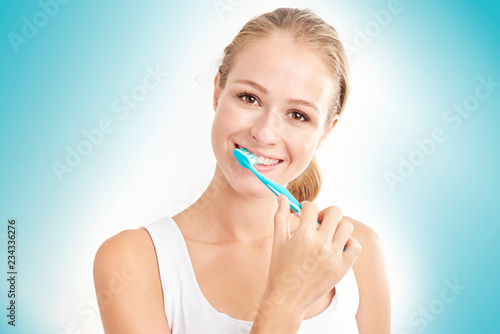 Studio shot of beautiful young woman brushing her teeth while looking at camera and smiling. Isolated on light blue background.