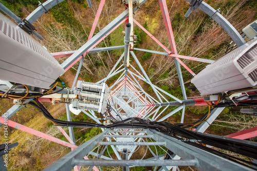 Red telecommunication tower or mast with microwave, radio panel antennas, outdoor remote radio units, power cables, coaxial cables, optic fibers are on the top mast that located in forest . View from