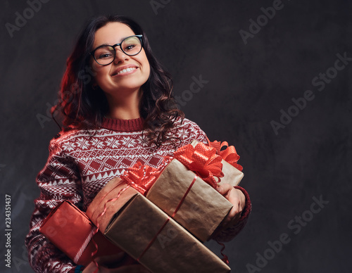 Portrait of a happy brunette girl wearing eyeglasses and warm sweater holding a gifts boxes, isolated on a dark textured background.