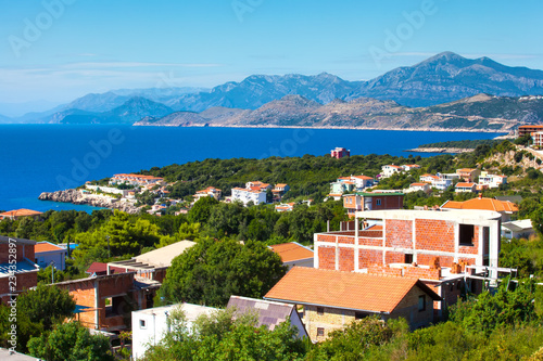 Brick houses on the beautiful green coast of the blue Adriatic Sea against the backdrop of magnificent mountains on the horizon on a sunny summer day. View from the highway, Ulcinj, Montenegro.