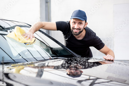 Handsome washer in t-shirt and cap wiping windshield with yellow microfiber at the open air car wash