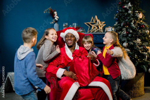 Children hugging African Santa Claus sitting on a red chair on the background of a Christmas tree.