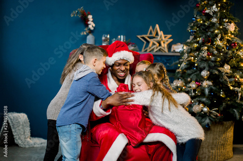Children hugging African Santa Claus sitting on a red chair on the background of a Christmas tree. © sofiko14