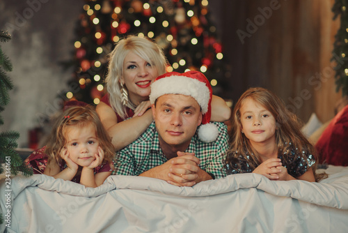 Merry Christmas and Happy Holidays! Cheerful parents and and two little children having fun and playing together near Christmas tree on the bed in living room