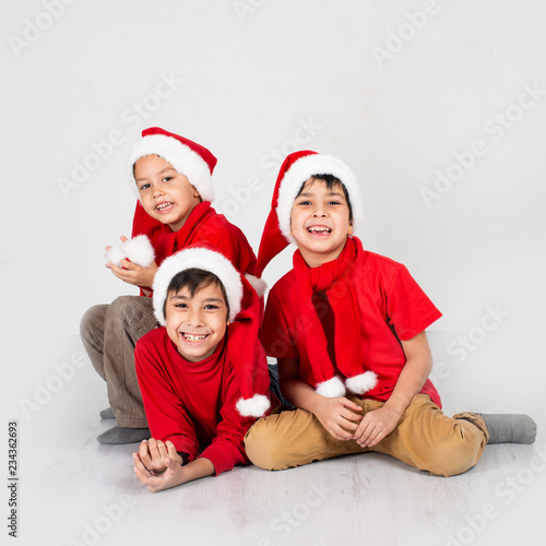 Full length portrait of a little  three boys wearing   red shirt and Santa Claus hats.  They sit together and smile in the studio