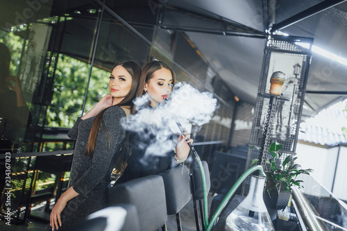 Two beautiful girls are standing on the summer terrace of a modern cafe and smoking hookah © Тарас Нагирняк