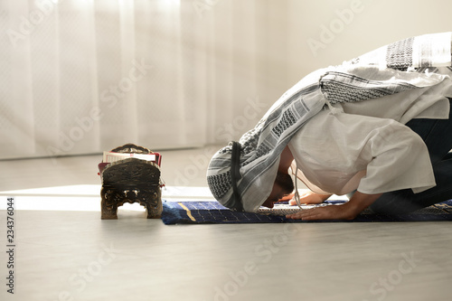 Muslim man in traditional clothes praying on rug indoors