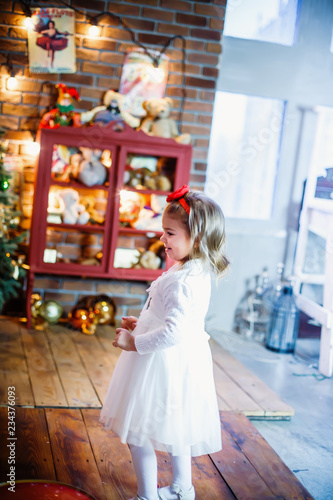little beautiful little girl in a white dress looking for toy near Christmas tree
