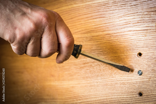 Screwdriver ready to turn flat head screw in finished board; handy man's hand and wood grain features.