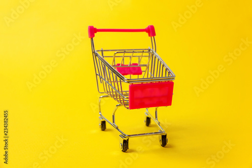 Small supermarket grocery push cart for shopping toy with wheels isolated on yellow colourful trendy modern fashion background. Sale buy mall market shop consumer concept. Copy space