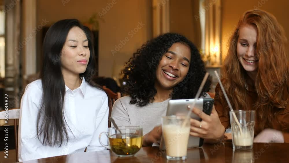 Multi Ethnic Group of Girls Laughing at a phone, a tablet Together In a Cafe Multi Ethnic Group of Girls Laughing at a phone, a tablet Together In a Cafe