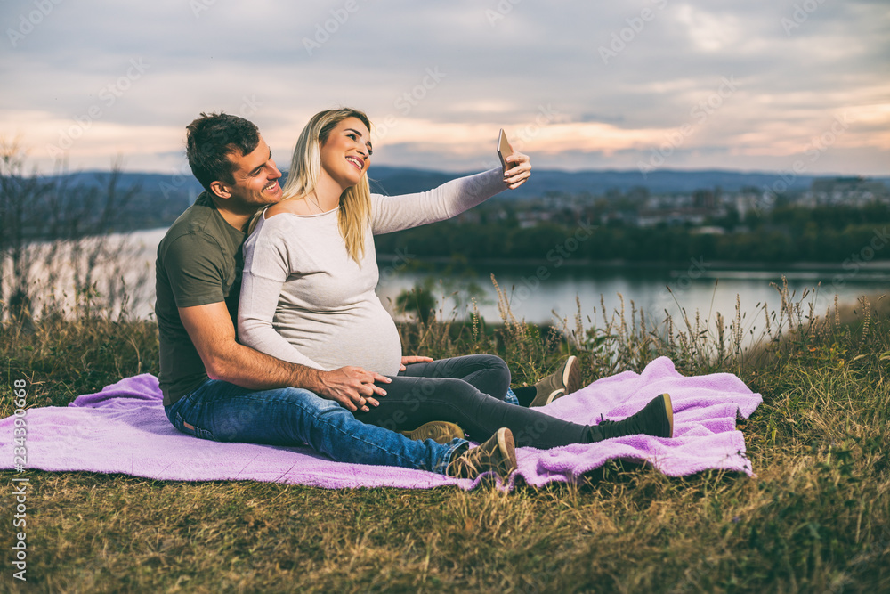 Happy husband and his pregnant wife taking selfie while they enjoy spending time together outdoor.