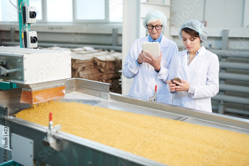 Waist up portrait of two female factory workers standing by macaroni conveyor belt during quality inspection at food production, copy space