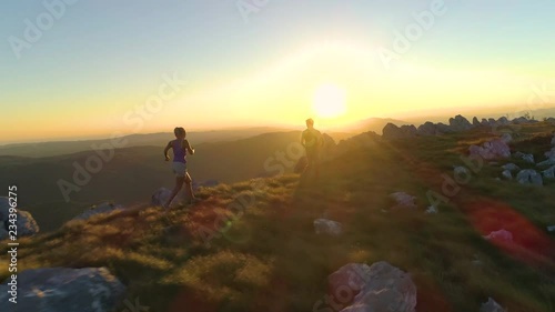 DRONE, SUN FLARE: Flying behind sporty tourist couple running along mountain trail at sunset. Golden morning sunbeams shine on athletic young man and woman training for a difficult fell running race.