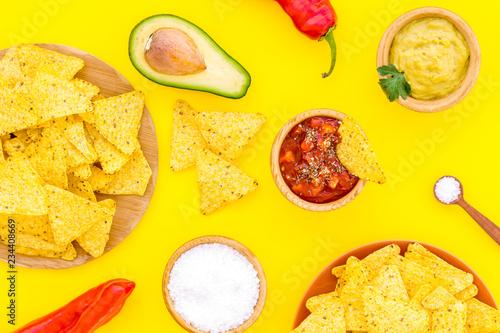 Popular mexican snack nachos. Tiangle nacho tortilla near salsa and guacamole sause  chili pepper  salt on yellow background top view
