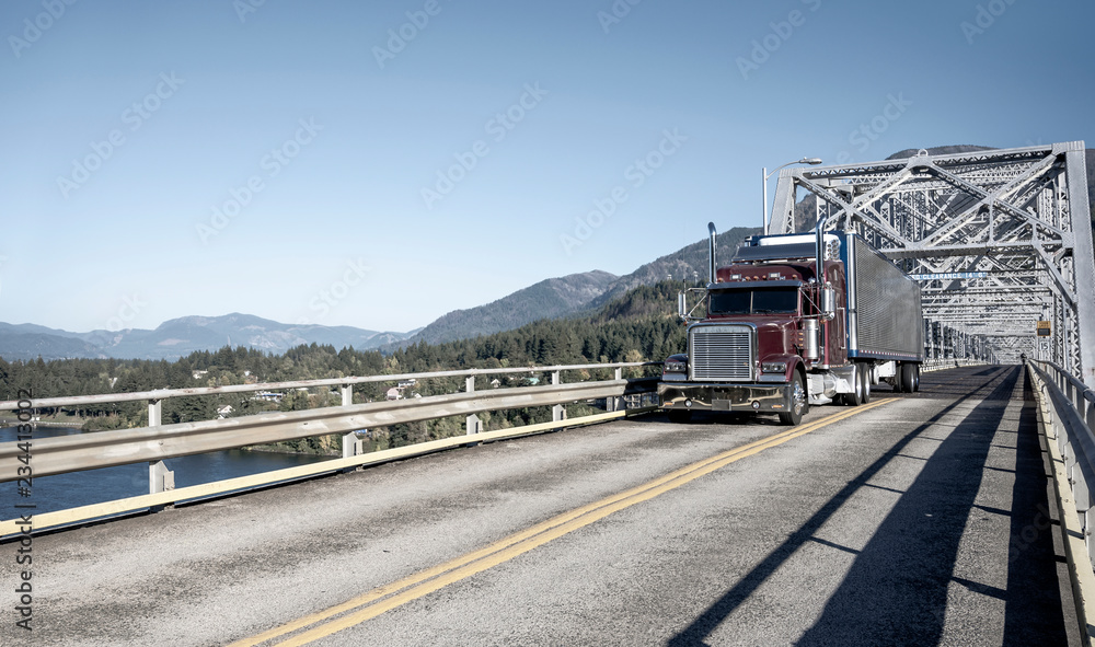 Big rig burgundy semi truck with shiny refrigerator semi trailer driving on the truss bridge in Columbia Gorge Big rig burgundy semi truck with shiny refrigerator semi trailer driving on the truss bridge in Columbia Gorge