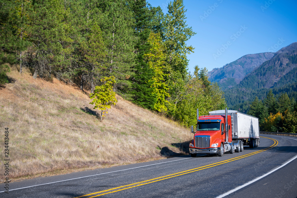 Big rig orange semi truck carry bulk semi trailer driving on winding road with trees and mountain