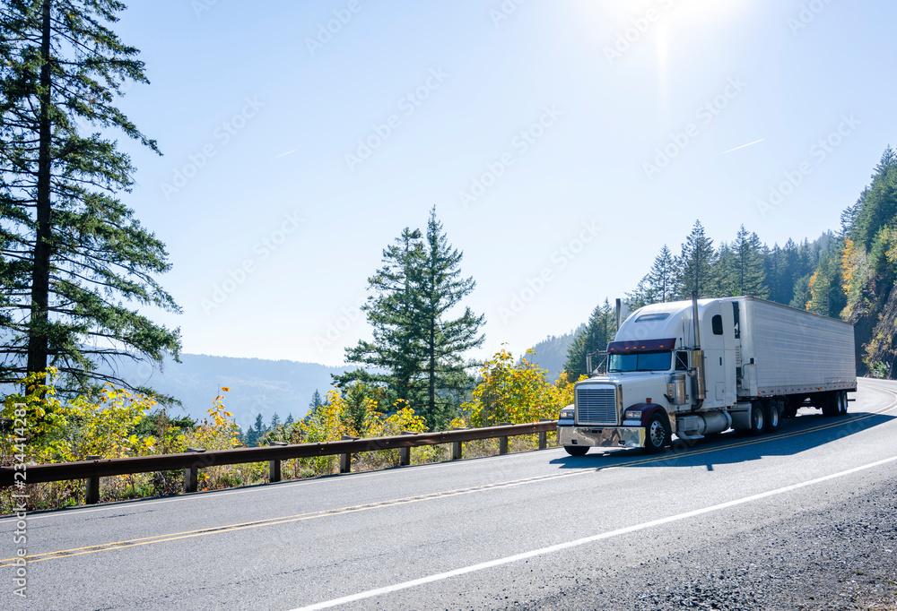 Big rig white classic semi truck transporting cargo in refrigerated semi trailer on winding autumn road