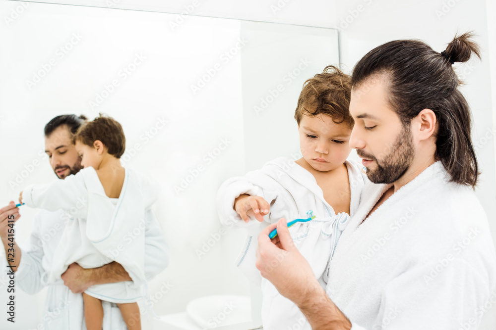 Toddler son looking at toothbrush with toothpaste and dad holding him in white bathroom Toddler son looking at toothbrush with toothpaste and dad holding him in white bathroom