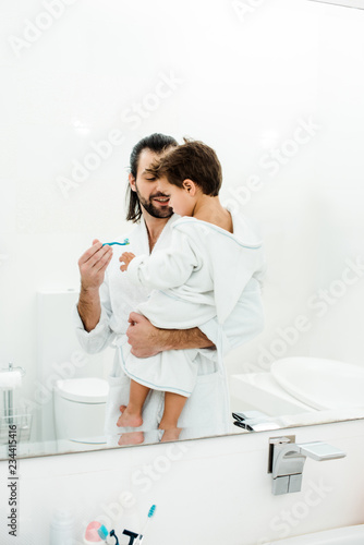 Father holding toddler son and showing toothbrush with toothpaste in white bathroom