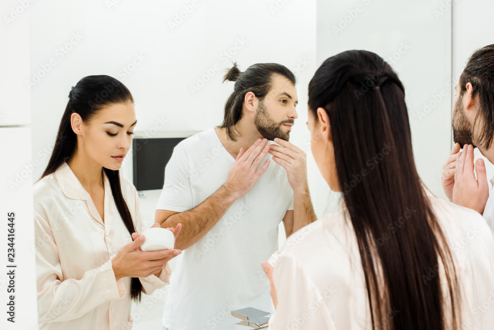 beautiful woman holding body cream while handsome man looking at mirror beautiful woman holding body cream while handsome man looking at mirror