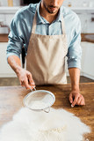 cropped shot of young man in apron sifting flour on kitchen table