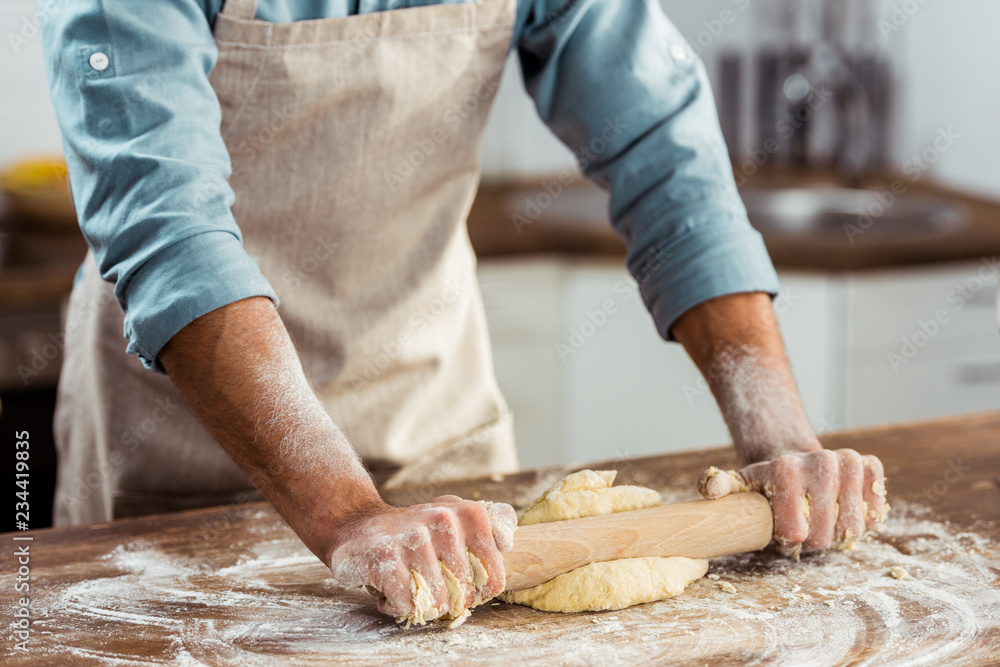 partial view of man in apron holding rolling pin and preparing dough in kitchen partial view of man in apron holding rolling pin and preparing dough in kitchen