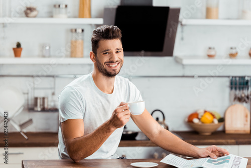 handsome young man holding cup of coffee and smiling at camera in kitchen