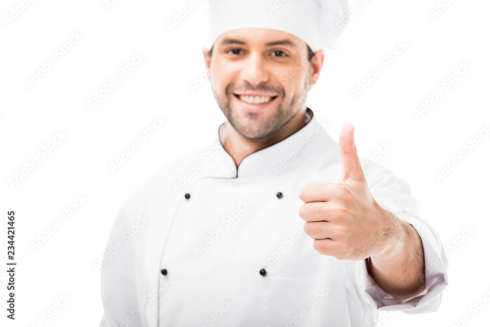 smiling young chef showing thumb up at camera isolated on white