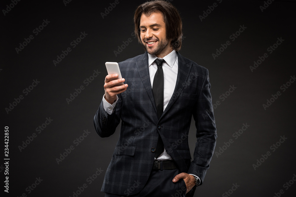 smiling young businessman standing with hand in pocket and using smartphone isolated on black smiling young businessman standing with hand in pocket and using smartphone isolated on black