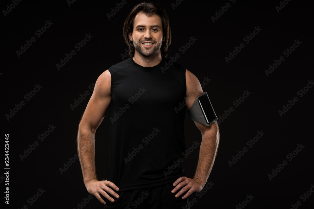 athletic young man with armband and smartphone standing with hands on waist and smiling at camera isolated on black athletic young man with armband and smartphone standing with hands on waist and smiling at camera isolated on black
