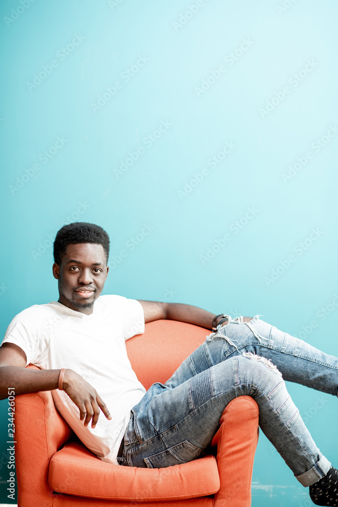 Portrait of a young african man dressed in white t-shirt and jeans sitting on the chair on the colorful background Portrait of a young african man dressed in white t-shirt and jeans sitting on the chair on the colorful background