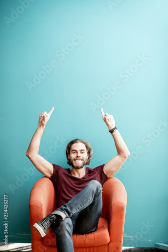 Portrait of a young caucasian bearded man with long hair showing with hands on the colorful background sitting on the chair. Image with copy space