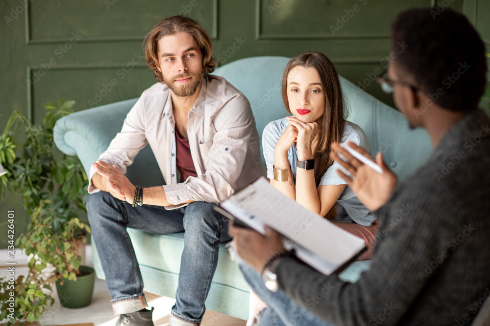 Young serious couple visiting a psychologist sitting on the comfortable couch during psychological session in the green office Young serious couple visiting a psychologist sitting on the comfortable couch during psychological session in the green office