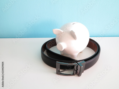 White ceramic piggy bank with black leather belt around on white table and blue wall behind