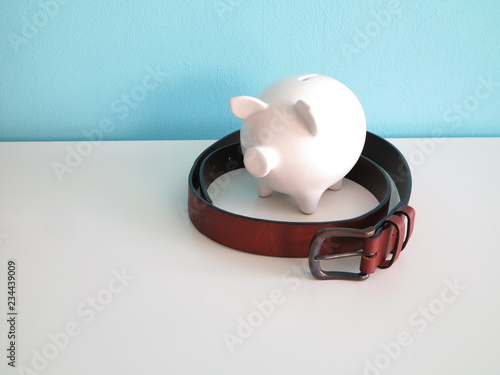 White ceramic piggy bank with brown leather belt around on white table and blue wall behind