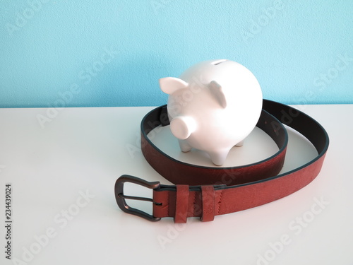 White ceramic piggy bank with brown leather belt around on white table and blue wall behind