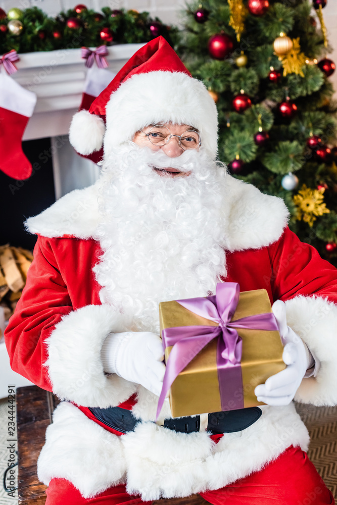 happy santa claus holding christmas present and looking at camera near christmas tree happy santa claus holding christmas present and looking at camera near christmas tree