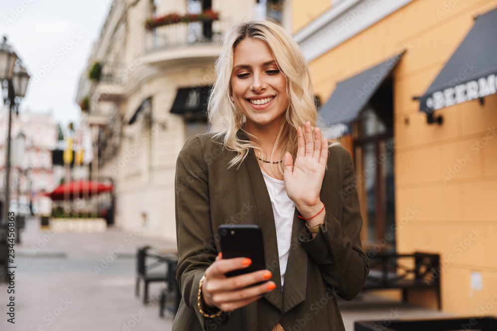 Image of attractive woman smiling and using mobile phone, while walking through city street Image of attractive woman smiling and using mobile phone, while walking through city street