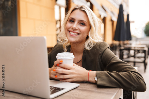 Portrait of lovely young woman holding takeaway coffee  and using silver laptop while sitting in cafe outdoor