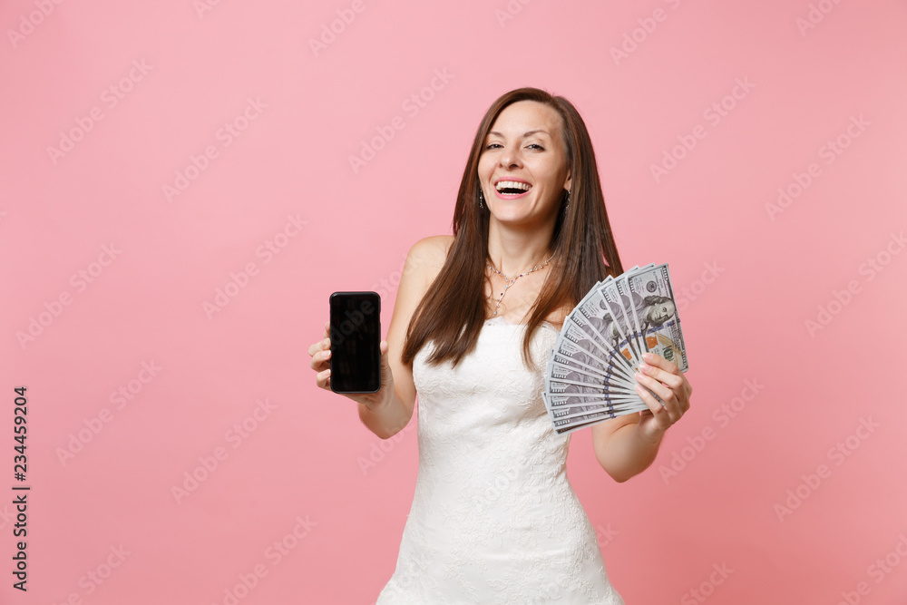 Joyful bride woman in wedding dress holding bundle lots of dollars, cash money, mobile phone with blank black empty screen isolated on pink background. Organization of wedding celebration. Copy space. Joyful bride woman in wedding dress holding bundle lots of dollars, cash money, mobile phone with blank black empty screen isolated on pink background. Organization of wedding celebration. Copy space.