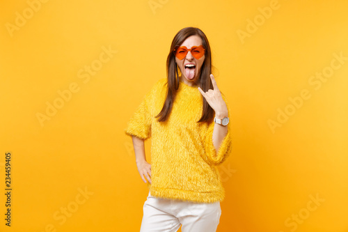 Funny crazy young woman in fur sweater and heart orange glasses screaming, showing rock-n-roll sign isolated on bright yellow background. People sincere emotions, lifestyle concept. Advertising area.