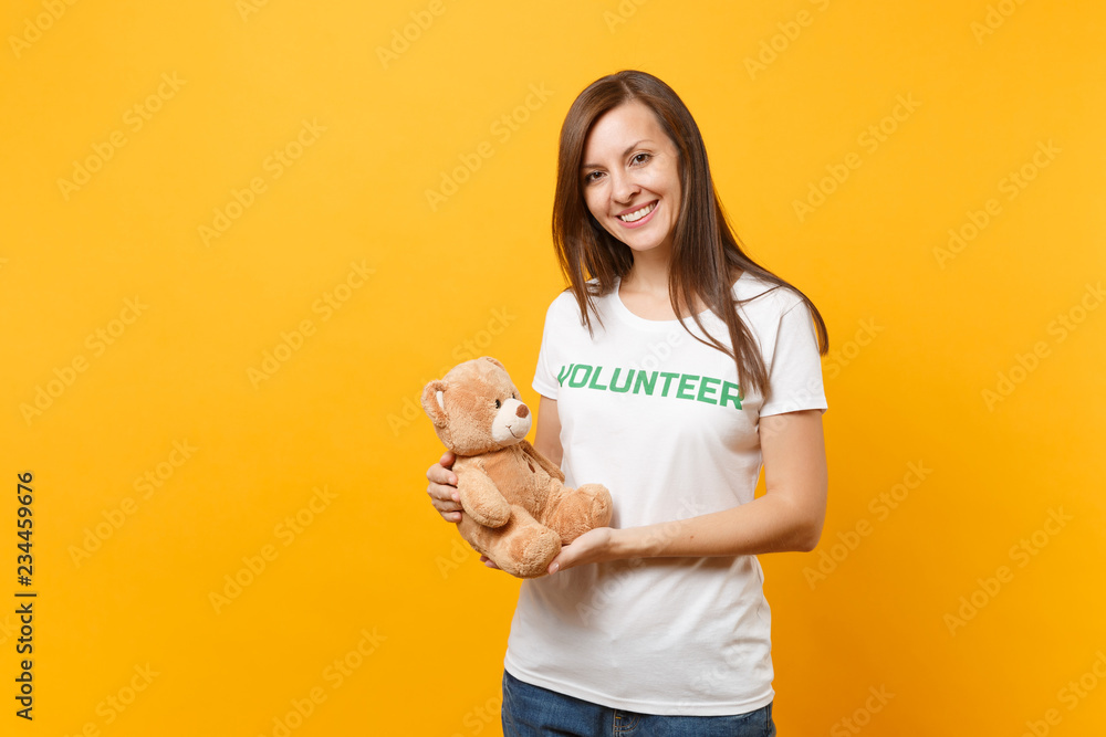 Portrait of woman in white t-shirt with written inscription green title volunteer hold teddy bear plush toy isolated on yellow background. Voluntary free assistance help, charity grace work concept. Portrait of woman in white t-shirt with written inscription green title volunteer hold teddy bear plush toy isolated on yellow background. Voluntary free assistance help, charity grace work concept.