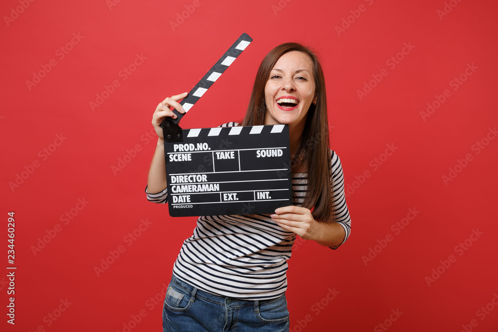 Φωτογραφία Portrait of laughing young woman in striped clothes holding classic black film making clapperboard isolated on bright red background
