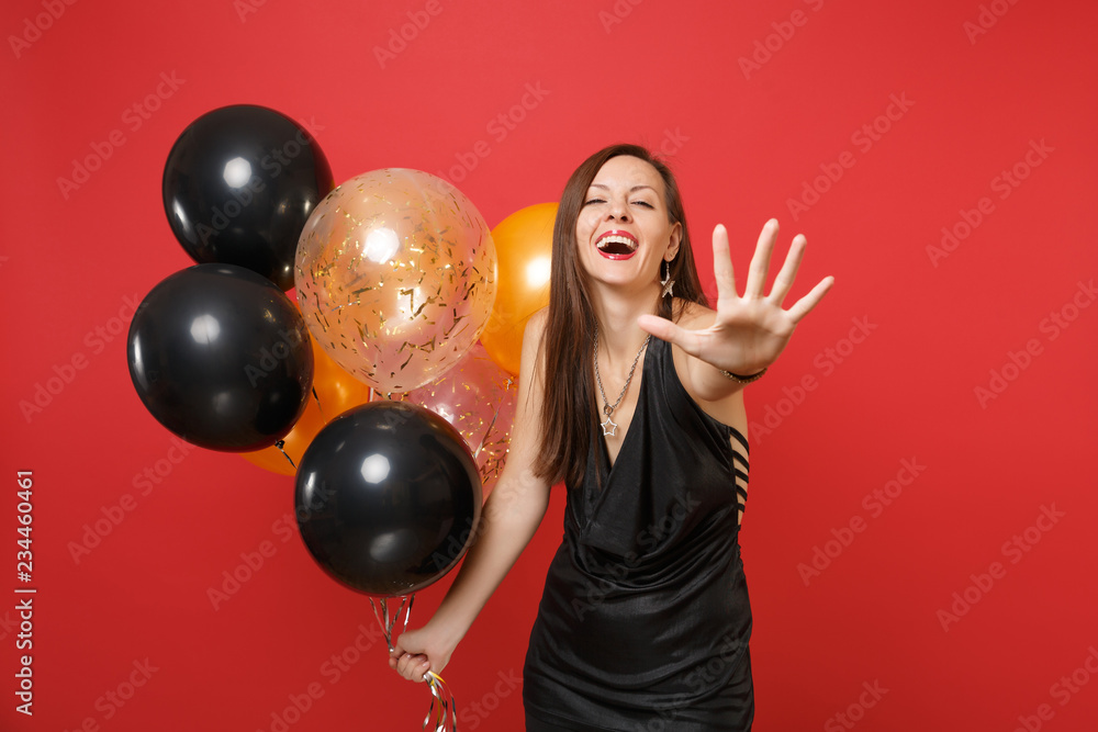 Fototapeta premium Laughing cheerful young woman in black dress pointing hand on camera and holding air balloons, celebrating isolated on red background. Women's Day Happy New Year birthday mockup holiday party concept.