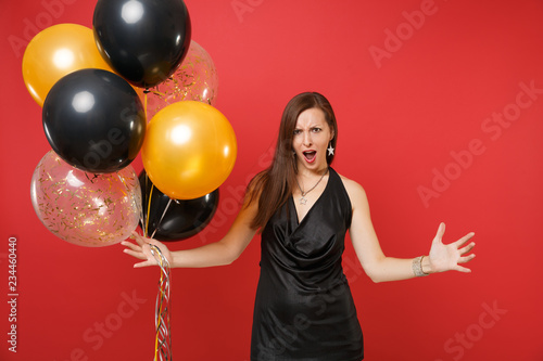 Irritated young woman in black dress celebrating swearing, spreading hands holding air balloons isolated on red background. St. Valentine's Day, Happy New Year, birthday mockup holiday party concept. © ViDi Studio