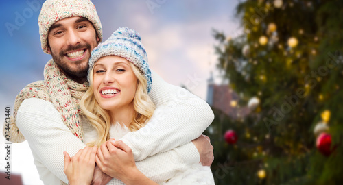 winter and holidays concept - happy couple in hats hugging over christmas tree at tallinn old town hall square background