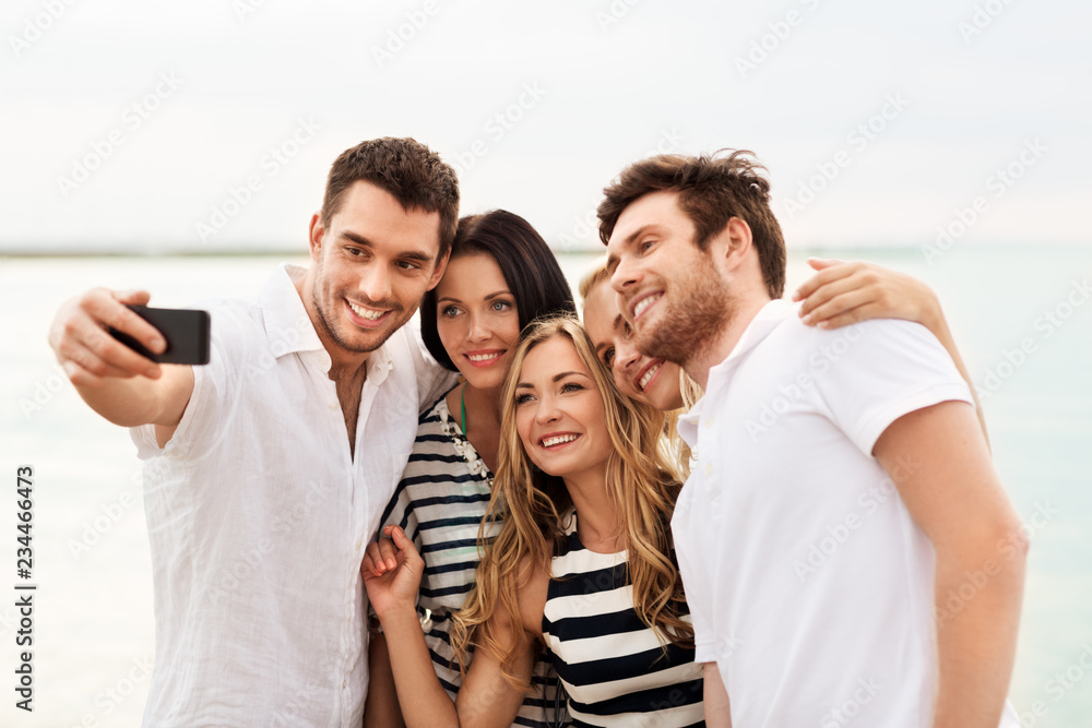 Obraz premium friendship, summer holidays and people concept - group of happy friends taking picture by smartphone on beach