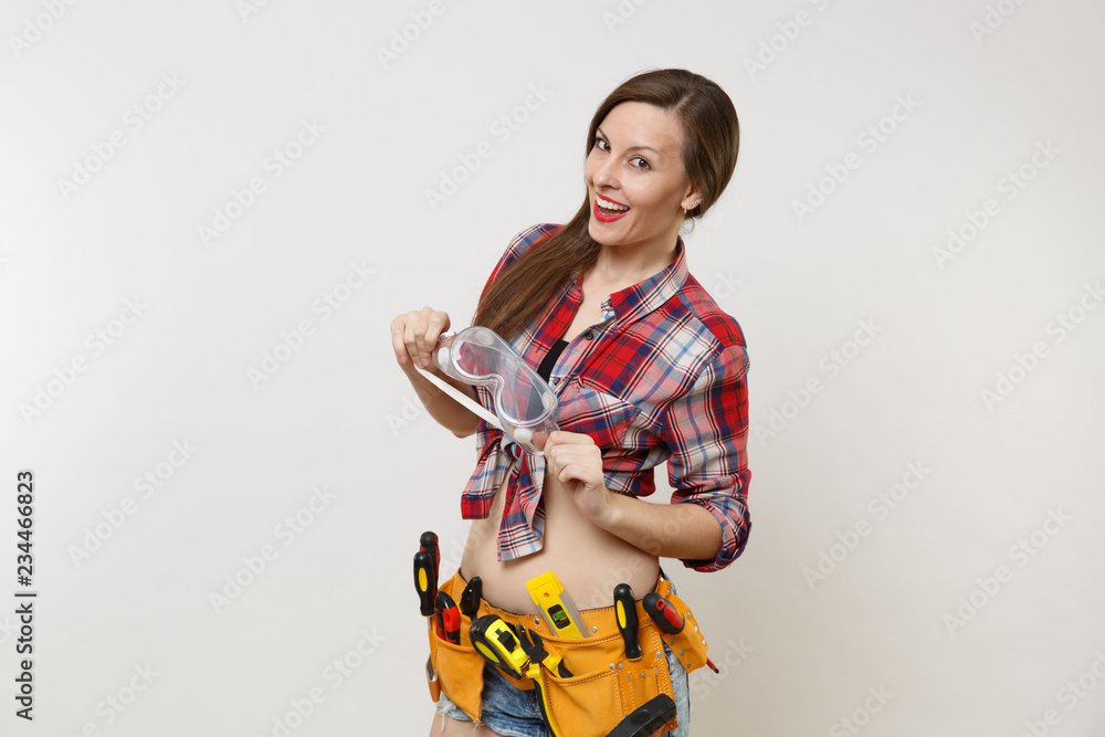 Strong young handyman woman in plaid top shirt, denim shorts, kit tools belt full of instruments in protective goggles isolated on white background. Female in male work. Copy space for advertisement Strong young handyman woman in plaid top shirt, denim shorts, kit tools belt full of instruments in protective goggles isolated on white background. Female in male work. Copy space for advertisement