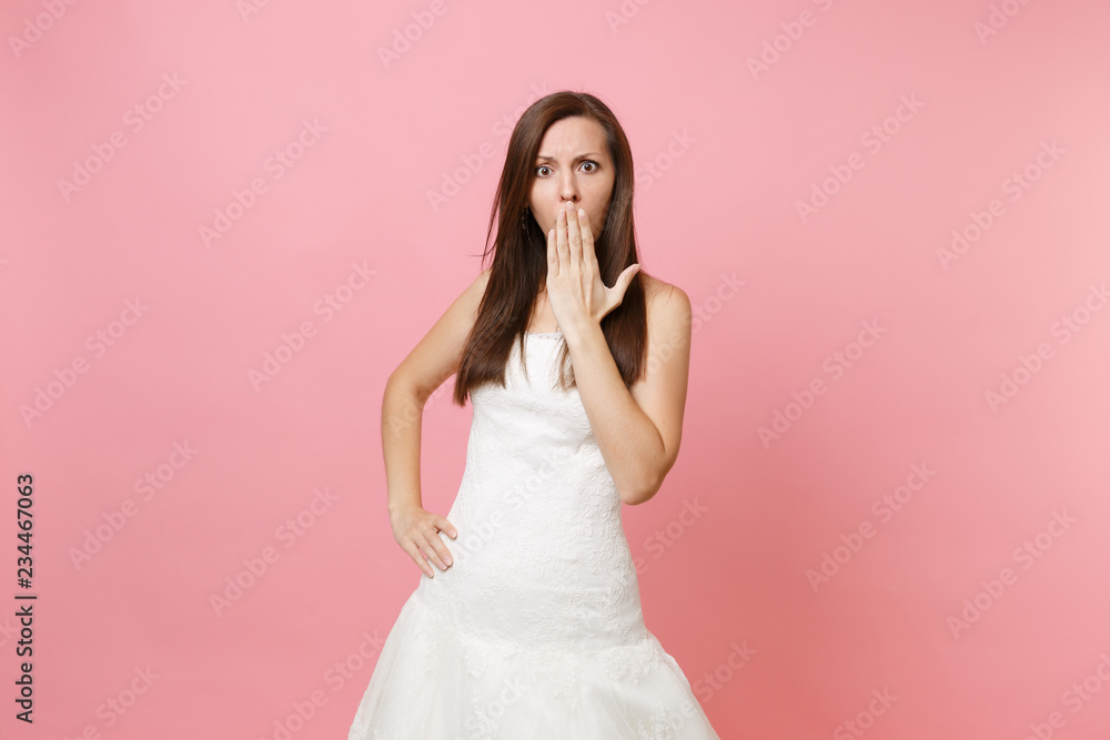 Portrait of concerned shocked bride woman in lace white wedding dress keeping hand on mouth covering face isolated on pastel pink background. Wedding celebration concept. Copy space for advertisement. Portrait of concerned shocked bride woman in lace white wedding dress keeping hand on mouth covering face isolated on pastel pink background. Wedding celebration concept. Copy space for advertisement.